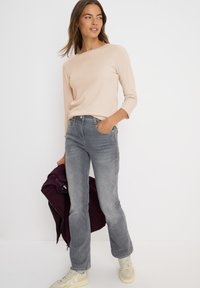 Haut beige à manches longues, jeans gris et baskets beiges. Le modèle tient une veste bordeaux. Tissu doux et coupe décontractée. Design simple.