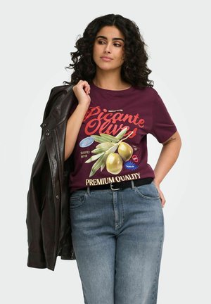 Kvinde med krøllet hår iført en mørkerød t-shirt med olivengrafik og tekst, holder en sort læderjakke over skulderen, iført blå jeans.