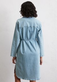 Robe en denim bleu clair avec des manches longues, une taille cintrée et des fentes sur les côtés. Elle présente un col et des détails de couture le long des coutures.