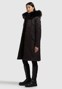 Musta toppatakki, jossa tekoeläinturkisvuorinen hupullinen, vetoketjullinen etuosa, sivutaskut ja pitkähkö siluetti, yhdistetty polven korkeudella oleviin mustiin saappaisiin.