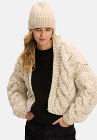 Kremowy, masywny kardigan z grubym splotem warkoczowym o krótkim fasonie, noszony na ciemnobrązowym, prążkowanym swetrze, dopełniony dopasowaną dzianinową czapką.