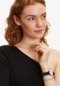 Zilveren ketting met onderling verbonden cirkels, gedragen over een geribbeld zwart topje; model houdt de ketting vast en toont een zilveren ring en een blauwe horloge.