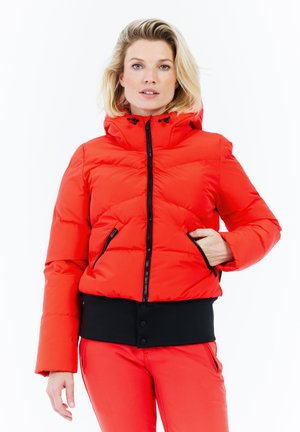 Blonde vrouw in een rode pufferjack met capuchon, zwarte taillepaneel en rode broek, staand tegen een witte achtergrond met hand in haar zak.