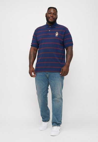 Marineblauwe gestreepte polo met een klein geborduurd berenlogo, gecombineerd met lichtblauwe jeans en witte sneakers. Korte mouwen en kraagontwerp.
