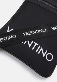 Pochette en nylon noir avec fermeture éclair, logo "VALENTINO" blanc bien visible et bandoulière réglable avec détail de logo.
