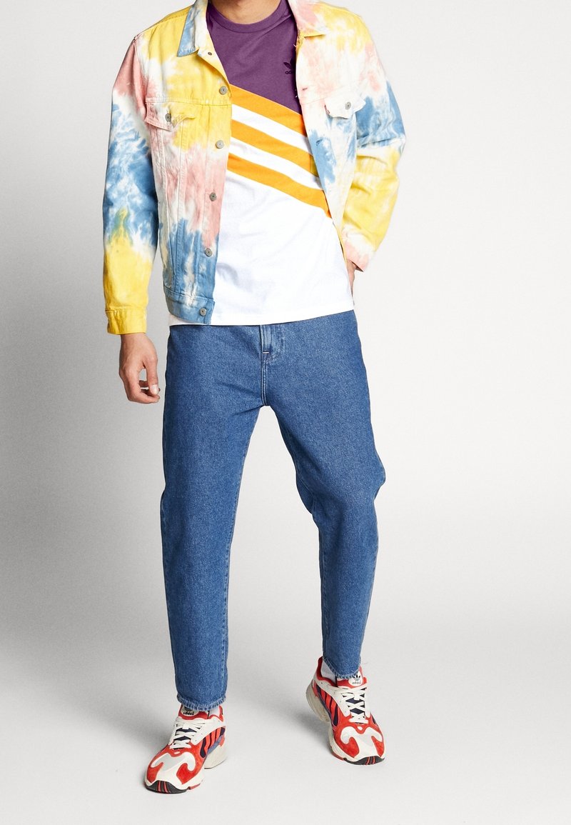 Täckjacka i pastellgul och blå batik, vit t-shirt med lila och orange ränder, löst sittande blå jeans och flerfärgade sneakers.