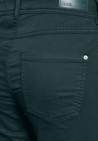 Tmavě zelené kalhoty s hladkým povrchem, které mají zadní kapsu se cvočkem a prošitý štítek na pase.