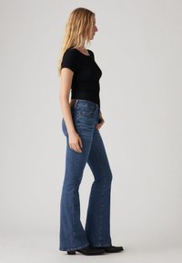 Mustade soonikuga lühikeste varrukatega topp, kõrge vöökohaga sinised lehviva säärega teksapüksid, sile pind, teksariie, mustad pahkluud saabad, külgvaade.