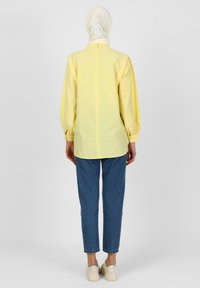 Blouse jaune à manches longues avec une fermeture éclair au dos, coupe décontractée et manches à poignet, associée à un jean droit bleu et des baskets blanches.