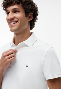 Young man with curly hair smiling while buttoning a white polo shirt with a small red, white, and blue logo on the chest.