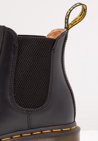 Black leather Chelsea boot featuring a textured mesh side panel, yellow stitching, and a pull tab with a contrasting yellow label.
