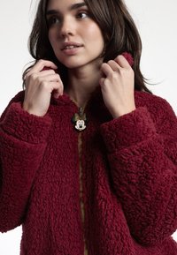 Chaqueta de forro polar burdeos con tejido texturizado, cremallera frontal y un charm de Minnie Mouse en el tirador de la cremallera. Cuenta con una capucha y puños anchos.