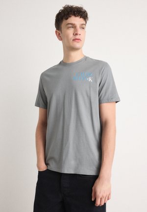 Gray cotton t-shirt featuring a crew neckline and short sleeves, with blue text logo on the left side. Paired with dark pants.
