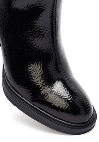 Botín negro de charol con una superficie lisa y brillante, punta redondeada y suela de goma resistente. Detalles de costura lateral visibles en la parte superior.
