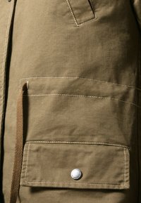 Olivgrön kanvasjacka med en framficka, silverfärgad tryckknapp och beige sömnadsdetaljer. Slät textur och praktisk design.