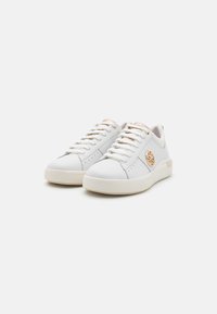 Sneaker bianche in pelle con finitura testurizzata, lacci piatti, perforazioni sulla punta e un logo circolare marrone chiaro sul lato.