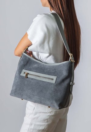 Femme vêtue de blanc portant un grand sac à bandoulière en suède gris avec une poche extérieure zippée sur son côté droit.