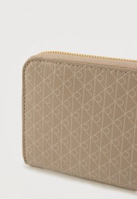 Μπεζ πορτοφόλι με απτική, διαμάντι μοτίβο που περιλαμβάνει καρδιές. Χρυσή φερμουάρ περιμετρικά, υφή υλικού και δομημένο σχήμα.