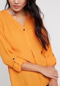 Orange blouse with a V-neck, rolled-up sleeves, and a black button detail. The fabric appears smooth with a slightly shiny texture.