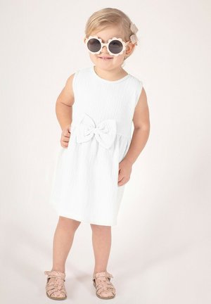 Biała sukienka bez rękawów z teksturowanej tkaniny z kokardą z przodu, zestawiona z różowymi sandałami w kwiaty i okrągłymi okularami przeciwsłonecznymi w kształcie kwiatów.