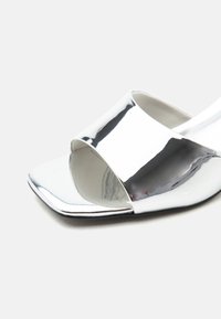 Λείτο ασημί σανδάλι ανοιχτού δακτύλου με φαρδιά λουρίδα και τετράγωνο σχέδιο δακτύλου σε απλό φωτεινό φόντο.