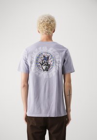 Camiseta de algodón de color púrpura claro con un diseño gráfico de una pata dentro de un círculo, rodeada de texto y patrones de llamas coloridas en la parte trasera.