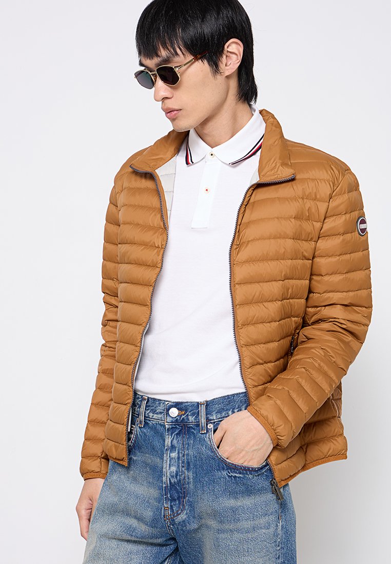 Giacca a vento color nocciola chiaro con cuciture orizzontali e chiusura a zip. Indossata su una polo bianca con colletto rosso e blu navy.