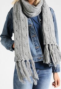 Écharpe en tricot gris avec un motif texturé et des extrémités effilochées, portée lâchement autour du cou, complétée par une veste en jean.