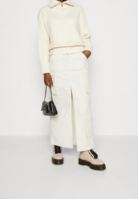 Krémový žebrovaný pulovr s polozipem, doplněný dlouhou bílou sukní s kapsami cargo. Celý outfit doplňuje černá kabelka a béžové šněrovací boty.