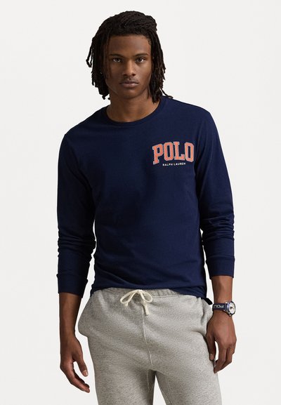 Mørkeblå langermet t-skjorte laget av bomull, med en rød og hvit "POLO" logo og rund halsdesign. Kombinert med grå joggebukser.