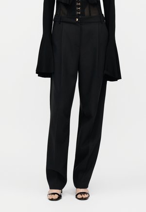 Άτομο που φοράει ψηλόμεσο μαύρο παντελόνι με πιέτες, συνδυασμένο με μαύρο μακρυμάνικο τοπ με φαρδιά μανίκια και μαύρα ανοιχτά πέδιλα.