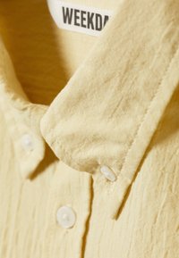 Camisa de manga corta en tejido amarillo claro. Cuenta con un collar clásico, dos botones blancos y un material texturizado. Etiqueta de la marca visible.