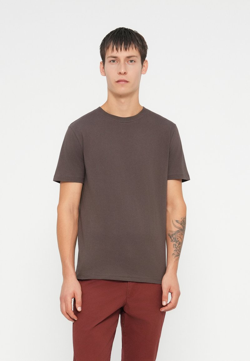 Camiseta de manga corta marrón oscuro con cuello redondo clásico, hecha de tela suave. Textura lisa, diseño minimalista, con un tatuaje visible en el brazo.