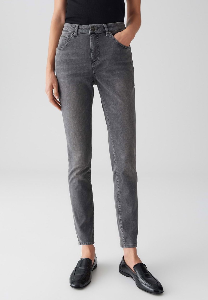 Donna che indossa jeans slim-fit grigi e mocassini neri, in piedi contro uno sfondo chiaro uniforme con una mano nella tasca.