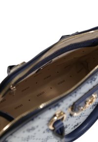 Tengerészkék és fehér mintás kézitáska sima texture-rel, arany színű fémrészletekkel és bézs belsővel, amelyen a "GUESS" logó ismétlődik.