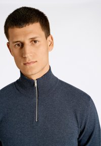 Tummansininen vetoketjullinen villapaita, joka on valmistettu pehmeästä neulemateriaalista, ja jossa on korkea kaulus sekä hopeinen vetoketju. Malli on yksinkertainen mutta moderni.