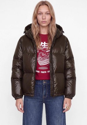 Modèle portant une doudoune à capuche marron foncé par-dessus un tee-shirt rouge à motif graphique et un jean bleu, sur un fond blanc uni.