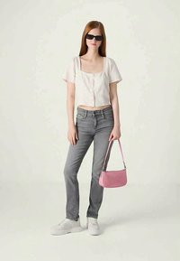 Balta cropped pogu krekla, pelēkas džinses, baltas kedas, rozā rokassomas un melni saulesbrilles; modele stāv uz gaiša fona.