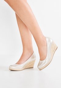 Des chaussures à talons compensés argent métallisé avec une tige synthétique lisse, un bout arrondi et un accent en jute tissé, reposant sur une semelle en caoutchouc beige.