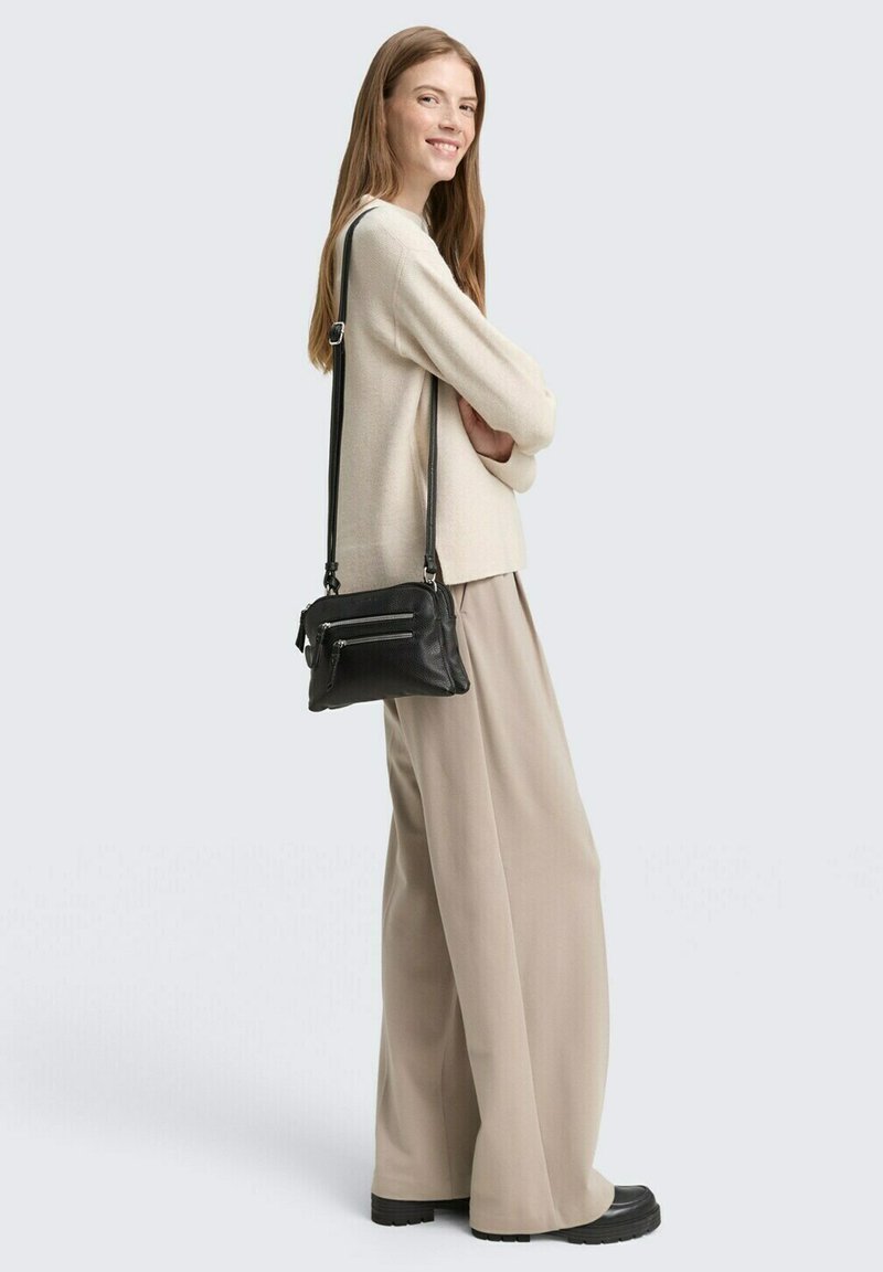 Schwarze Leder-Crossbody-Tasche mit zwei vorderen Reißverschlussfächern, ergänzt durch einen langen verstellbaren Gurt. Das Modell trägt einen beigen Pullover und weite beige Hosen.