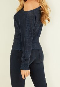 Granatowy sweter z odkrytymi ramionami, wykonany z przędzy metalizowanej, z długimi rękawami i fakturowanym materiałem, dopasowany w talii, zestawiony z ciemnymi dżinsami.