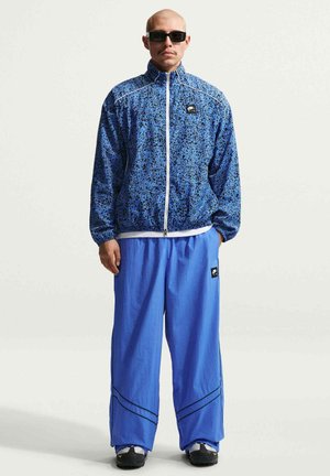 Man met zwarte zonnebril, een blauwe gebloemde jas met rits, losse effen blauwe broek en zwart-witte sneakers, staand tegen een witte achtergrond.