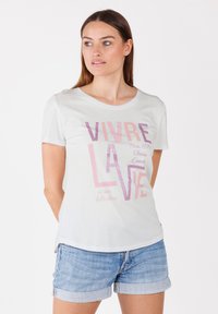 T-shirt gris clair à manches courtes avec un col rond, ornée de texte rose et pailleté. Assortie à un short en jean, affichant une coupe décontractée.