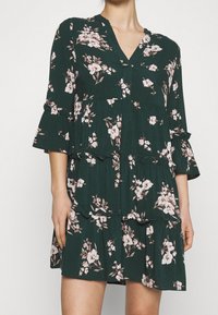 Blommig klänning i mörkgrön med rosa blommor. Har V-ringning, trekvartslånga ärmar med volanger och en skuren kjoldesign.