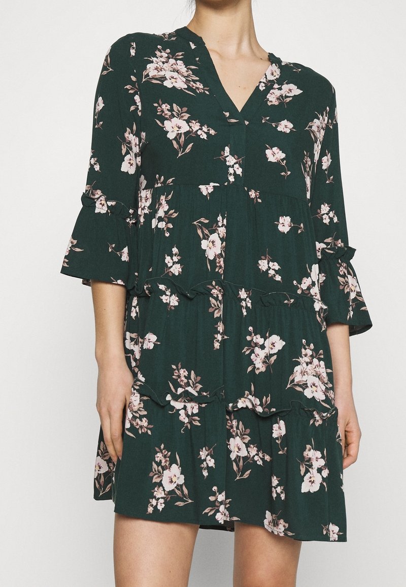 Blommig klänning i mörkgrön med rosa blommor. Har V-ringning, trekvartslånga ärmar med volanger och en skuren kjoldesign.