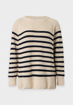 Kvinde iført beige og sort stribet sweater, mørkebrun lædernederdel med frontslids, sorte støvler, holder en sort håndtaske.