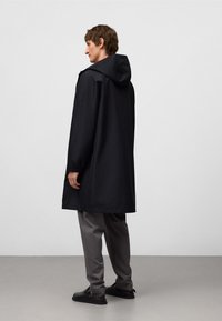 Svart vattentät jacka med huva, med avslappnad passform och långa ärmar, bärs över grå byxor och svarta skor. Enkel design.