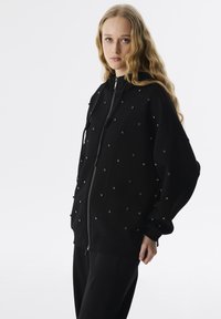 Felpa nera con zip realizzata in un tessuto morbido, caratterizzata da decorazioni circolari su tutta la superficie. Progettata con cappuccio e polsini elasticizzati.