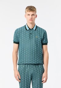 Pikétröja i teal med geometriskt mönster. Har en kontrasterande svart krage med blå och vita ränder. Korta ärmar och Lacoste-logotyp.