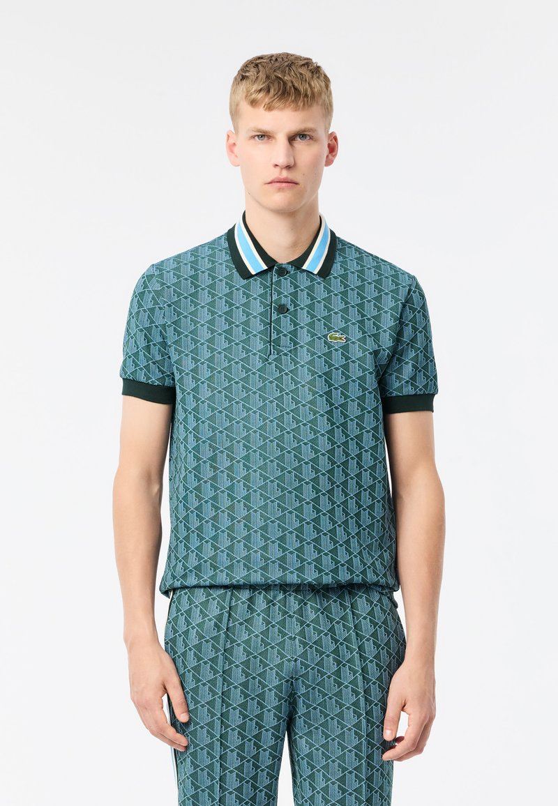 Pikétröja i teal med geometriskt mönster. Har en kontrasterande svart krage med blå och vita ränder. Korta ärmar och Lacoste-logotyp.